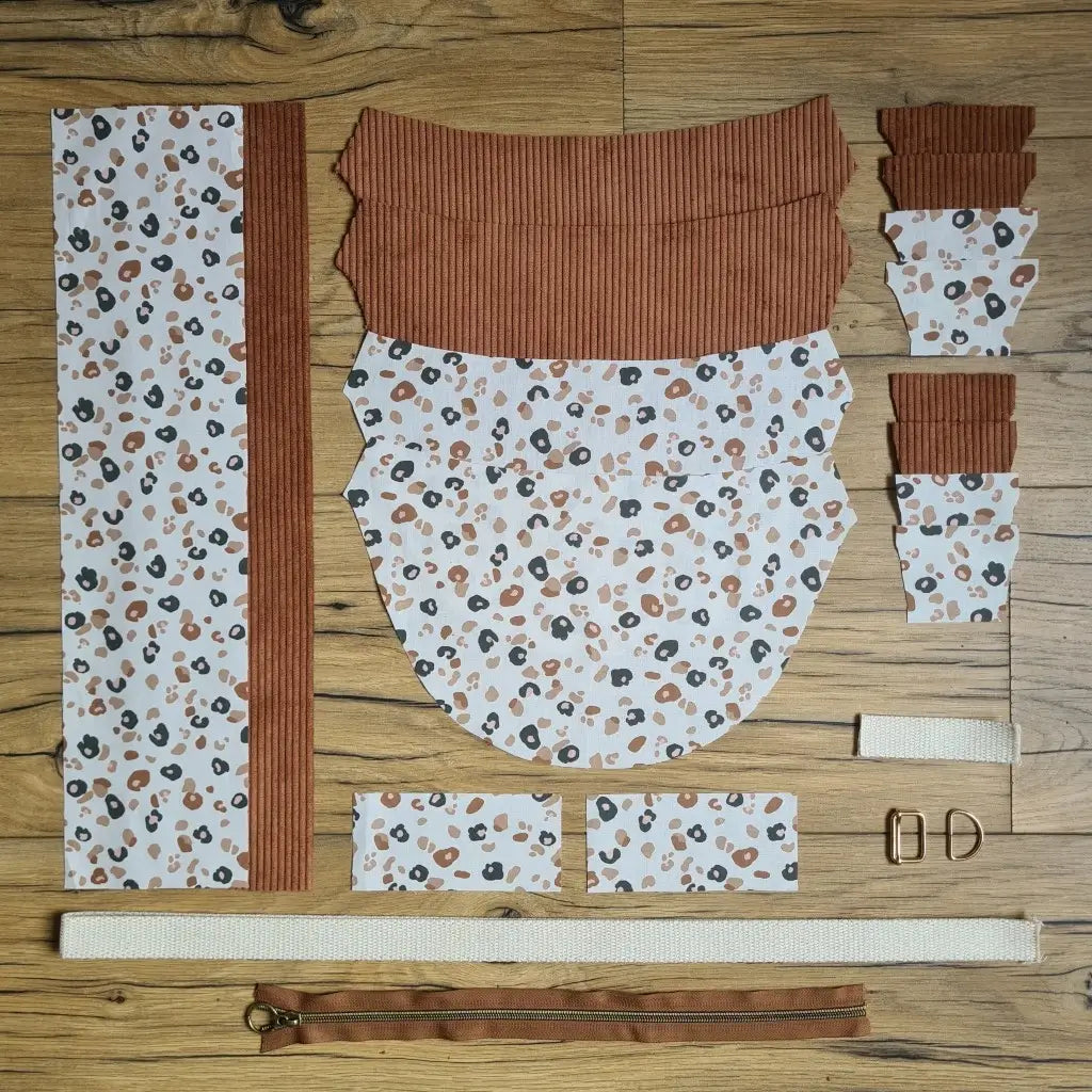 Kit "prêt à coudre" pour un sac demi-lune en velours côtelé au coloris marron avec une doublure en tissu à motif léopard. L'ensemble des pièces de tissu, déjà prédécoupées aux bonnes dimensions, sont disposées au sol et comprennent neuf pièces en tissu à motif léopard et sept pièces en velours côtelé de couleur marron ainsi qu'une fermeture éclair marron et une sangle beige.