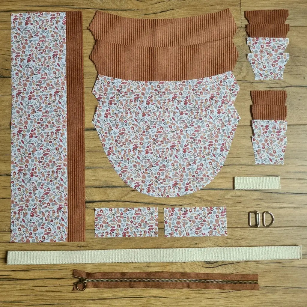 Kit "prêt à coudre" pour un sac demi-lune en velours côtelé au coloris marron avec une doublure en tissu à motif liberty. L'ensemble des pièces de tissu, déjà prédécoupées aux bonnes dimensions, sont disposées au sol et comprennent neuf pièces en tissu à motif liberty et sept pièces en velours côtelé de couleur marron ainsi qu'une fermeture éclair marron et une sangle beige.