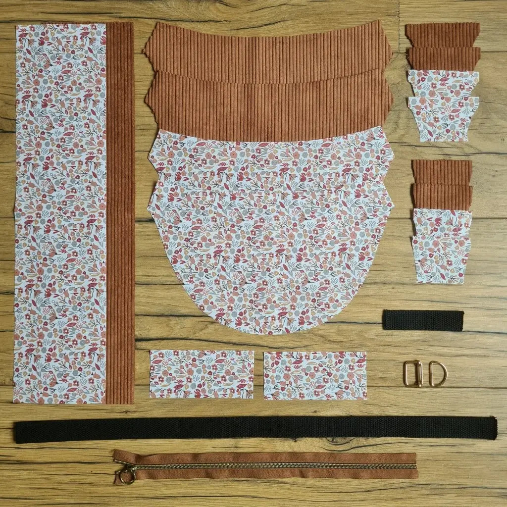 Kit "prêt à coudre" pour un sac demi-lune en velours côtelé au coloris marron avec une doublure en tissu à motif liberty. L'ensemble des pièces de tissu, déjà prédécoupées aux bonnes dimensions, sont disposées au sol et comprennent neuf pièces en tissu à motif liberty et sept pièces en velours côtelé de couleur marron ainsi qu'une fermeture éclair marron et une sangle noire.