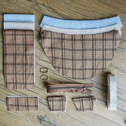 Ensemble de pièces prédécoupées pour confectionner un sac demi-lune, incluant tissu à carreaux beige et noir, bandes assorties, doublure claire, sangle en coton écru, fermeture éclair marron et accessoires métalliques dorés, disposés sur parquet. Ce kit créatif complet permet de réaliser facilement un sac chic et moderne grâce à un assemblage couture simple.