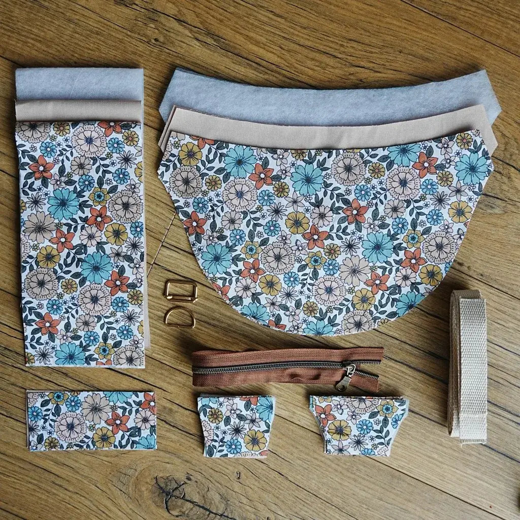 Kit complet pour créer un sac demi-lune comprenant tissu en coton imprimé fleuri style rétro aux tons bleu, orange et moutarde, doublure beige, renfort en ouate, sangle en coton écru, fermeture éclair marron et boucles métalliques dorées, disposés sur un sol en bois. Idéal pour un projet couture DIY alliant originalité et élégance.