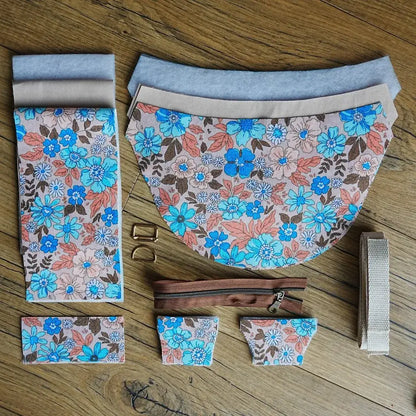 Ensemble de pièces prédécoupées pour confectionner un sac demi-lune, avec tissu en coton à grands motifs floraux bleu vif et orangé, doublure unie claire, sangle écrue, zip marron et accessoires métalliques dorés. Disposé à plat sur un sol en bois, ce kit couture complet permet de réaliser facilement un sac unique et éclatant.
