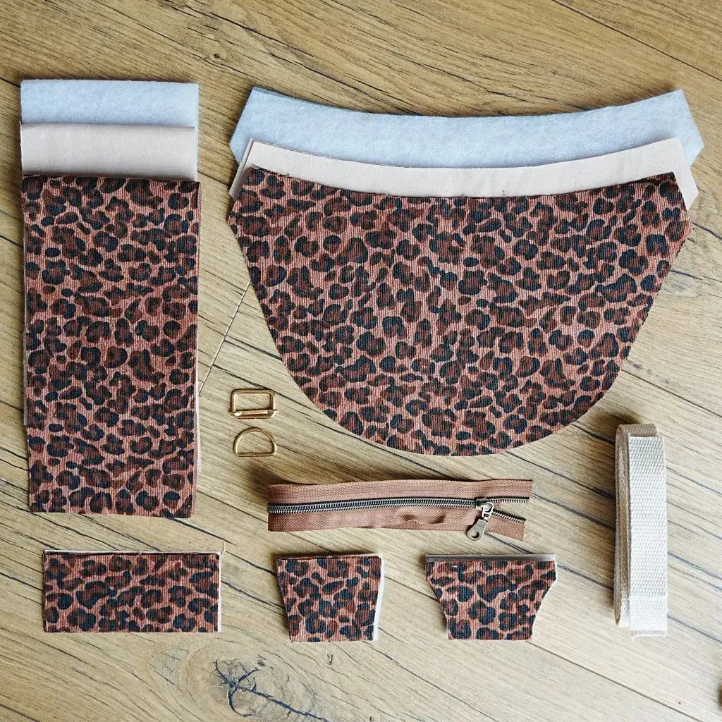 Kit de couture pour sac demi-lune comprenant tissu imprimé motif léopard marron et noir, doublure unie beige, renfort en ouate, sangle en coton écru, fermeture éclair marron et boucles métalliques dorées. Les éléments sont disposés à plat sur un sol en bois. Idéal pour un projet DIY original permettant de réaliser un sac tendance au style sauvage et affirmé.