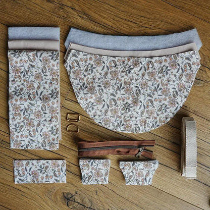 Kit prêt à assembler pour sac demi-lune, composé de pièces en coton imprimé fleuri, doublure claire, renfort en ouate, sangle en coton écru, fermeture éclair marron et boucles métalliques dorées. Disposé à plat sur un sol en bois, cet ensemble de couture DIY permet de créer un sac élégant et délicat.