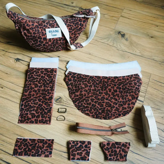 Kit DIY pour réaliser un sac banane demi-lune motif léopard, comprenant un modèle cousu en exemple et toutes les pièces nécessaires : tissu imprimé, doublure en ouate, sangle en coton écru, fermeture éclair marron et boucles métalliques dorées. Disposé sur un sol en bois, cet ensemble complet permet de confectionner un accessoire pratique et tendance.