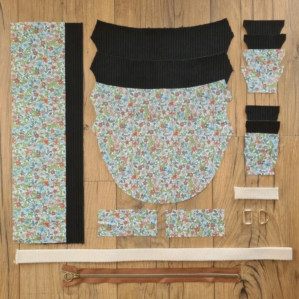 Kit "prêt à coudre" pour un sac demi-lune en velours côtelé au coloris noir avec une doublure en tissu à motif fleuri. L'ensemble des pièces de tissu, déjà prédécoupées aux bonnes dimensions, sont disposées au sol et comprennent neuf pièces en tissu à motif fleuri et sept pièces en velours côtelé de couleur noire ainsi qu'une fermeture éclair marron et une sangle blanche.