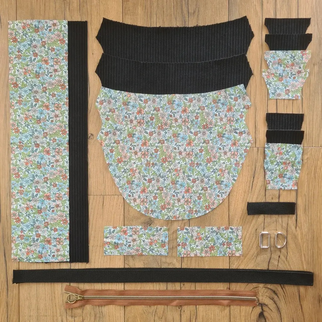 Kit "prêt à coudre" pour un sac demi-lune en velours côtelé au coloris noir avec une doublure en tissu à motif fleuri. L'ensemble des pièces de tissu, déjà prédécoupées aux bonnes dimensions, sont disposées au sol et comprennent neuf pièces en tissu à motif fleuri et sept pièces en velours côtelé de couleur noire ainsi qu'une fermeture éclair marron et une sangle noire.