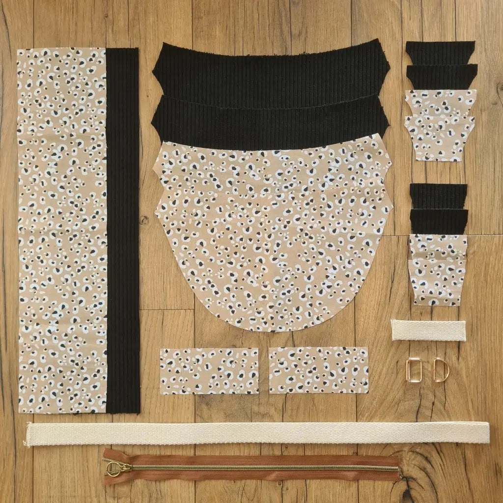 Kit "prêt à coudre" pour un sac demi-lune en velours côtelé au coloris noir avec une doublure en tissu à motif léopard. L'ensemble des pièces de tissu, déjà prédécoupées aux bonnes dimensions, sont disposées au sol et comprennent neuf pièces en tissu à motif léopard et sept pièces en velours côtelé de couleur noire ainsi qu'une fermeture éclair marron et une sangle blanche.