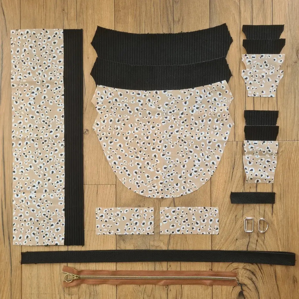 Kit "prêt à coudre" pour un sac demi-lune en velours côtelé au coloris noir avec une doublure en tissu à motif léopard. L'ensemble des pièces de tissu, déjà prédécoupées aux bonnes dimensions, sont disposées au sol et comprennent neuf pièces en tissu à motif léopard et sept pièces en velours côtelé de couleur noire ainsi qu'une fermeture éclair marron et une sangle noire.