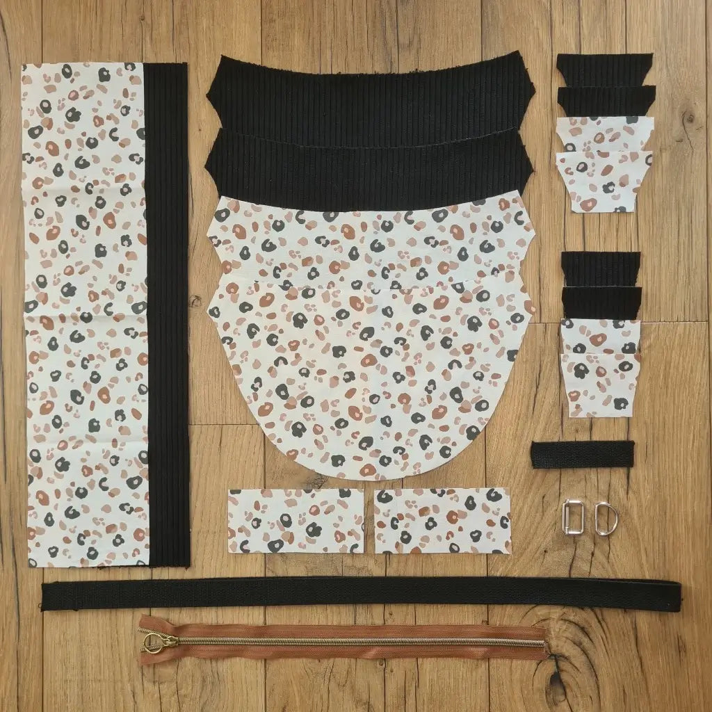 Kit "prêt à coudre" pour un sac demi-lune en velours côtelé au coloris noir avec une doublure en tissu à motif léopard. L'ensemble des pièces de tissu, déjà prédécoupées aux bonnes dimensions, sont disposées au sol et comprennent neuf pièces en tissu à motif léopard et sept pièces en velours côtelé de couleur noire ainsi qu'une fermeture éclair marron et une sangle noire.