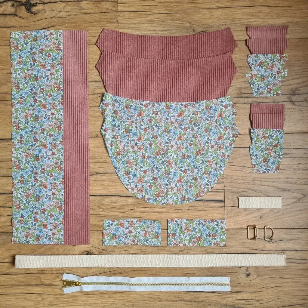 Kit "prêt à coudre" pour un sac demi-lune en velours côtelé au coloris rose avec une doublure en tissu à motif fleuri. L'ensemble des pièces de tissu, déjà prédécoupées aux bonnes dimensions, sont disposées au sol et comprennent neuf pièces en tissu à motif fleuri et sept pièces en velours côtelé de couleur rose ainsi qu'une fermeture éclair blanche et une sangle beige.