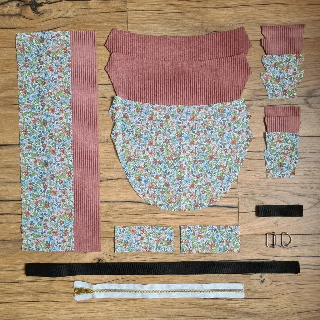 Kit "prêt à coudre" pour un sac demi-lune en velours côtelé au coloris rose avec une doublure en tissu à motif fleuri. L'ensemble des pièces de tissu, déjà prédécoupées aux bonnes dimensions, sont disposées au sol et comprennent neuf pièces en tissu à motif fleuri et sept pièces en velours côtelé de couleur rose ainsi qu'une fermeture éclair blanche et une sangle noire.