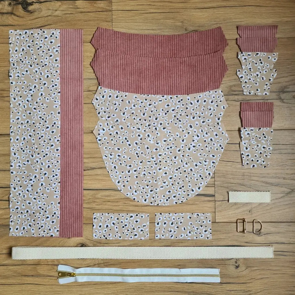 Kit "prêt à coudre" pour un sac demi-lune en velours côtelé au coloris rose avec une doublure en tissu à motif léopard. L'ensemble des pièces de tissu, déjà prédécoupées aux bonnes dimensions, sont disposées au sol et comprennent neuf pièces en tissu à motif léopard et sept pièces en velours côtelé de couleur rose ainsi qu'une fermeture éclair blanche et une sangle beige.