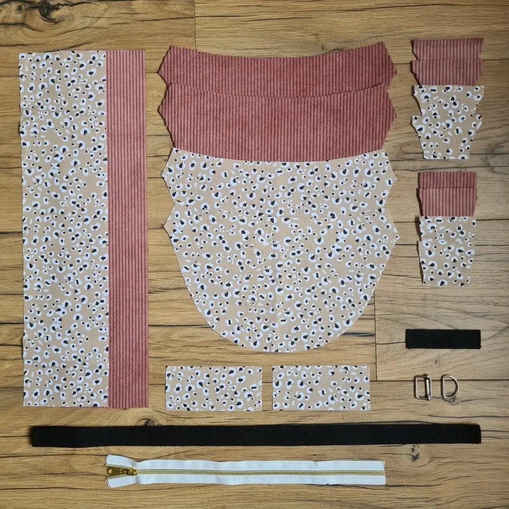 Kit "prêt à coudre" pour un sac demi-lune en velours côtelé au coloris rose avec une doublure en tissu à motif léopard. L'ensemble des pièces de tissu, déjà prédécoupées aux bonnes dimensions, sont disposées au sol et comprennent neuf pièces en tissu à motif léopard et sept pièces en velours côtelé de couleur rose ainsi qu'une fermeture éclair blanche et une sangle noire.