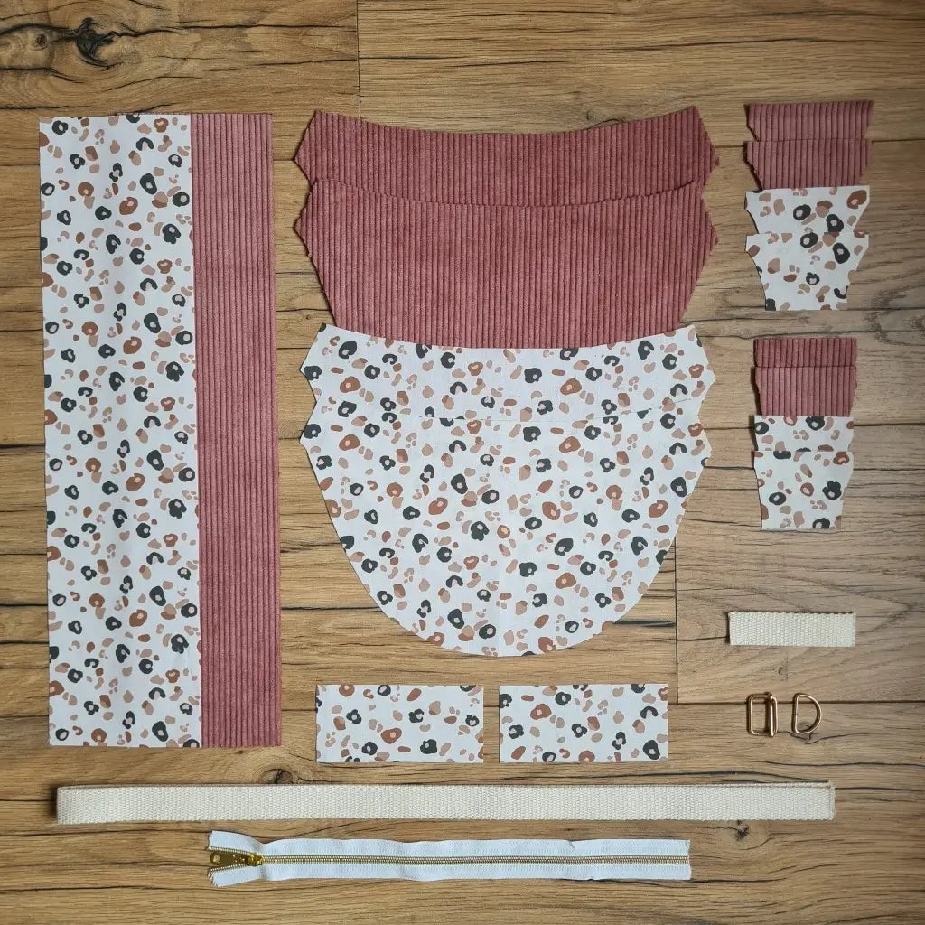 Kit "prêt à coudre" pour un sac demi-lune en velours côtelé au coloris rose avec une doublure en tissu à motif léopard. L'ensemble des pièces de tissu, déjà prédécoupées aux bonnes dimensions, sont disposées au sol et comprennent neuf pièces en tissu à motif léopard et sept pièces en velours côtelé de couleur rose ainsi qu'une fermeture éclair blanche et une sangle beige.