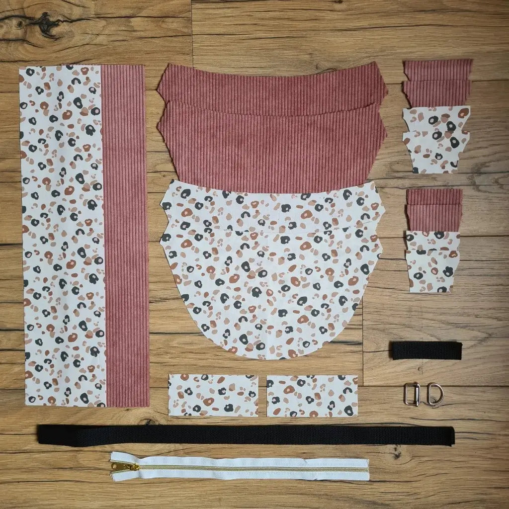 Kit "prêt à coudre" pour un sac demi-lune en velours côtelé au coloris rose avec une doublure en tissu à motif léopard. L'ensemble des pièces de tissu, déjà prédécoupées aux bonnes dimensions, sont disposées au sol et comprennent neuf pièces en tissu à motif léopard et sept pièces en velours côtelé de couleur rose ainsi qu'une fermeture éclair marron et une sangle noire.