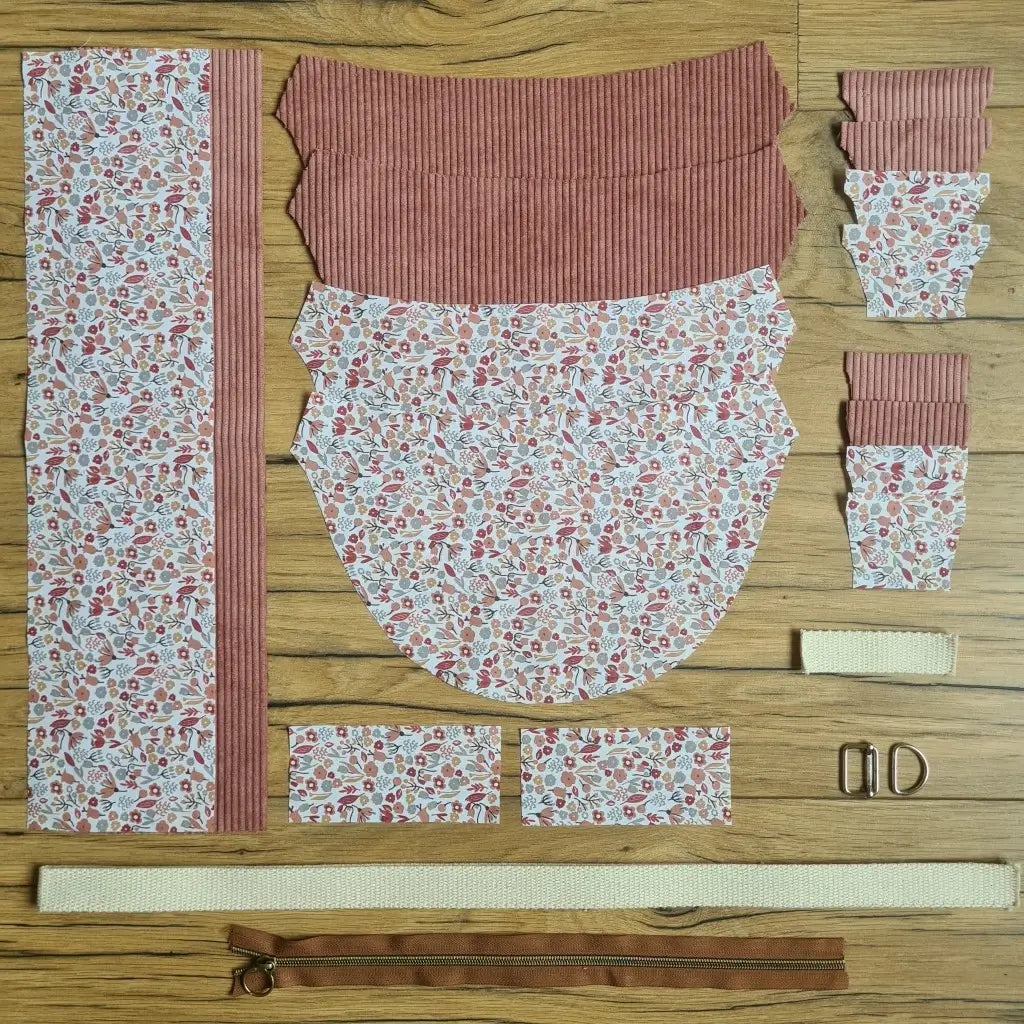 Kit "prêt à coudre" pour un sac demi-lune en velours côtelé au coloris rose avec une doublure en tissu à motif liberty. L'ensemble des pièces de tissu, déjà prédécoupées aux bonnes dimensions, sont disposées au sol et comprennent neuf pièces en tissu à motif liberty et sept pièces en velours côtelé de couleur rose ainsi qu'une fermeture éclair marron et une sangle beige.