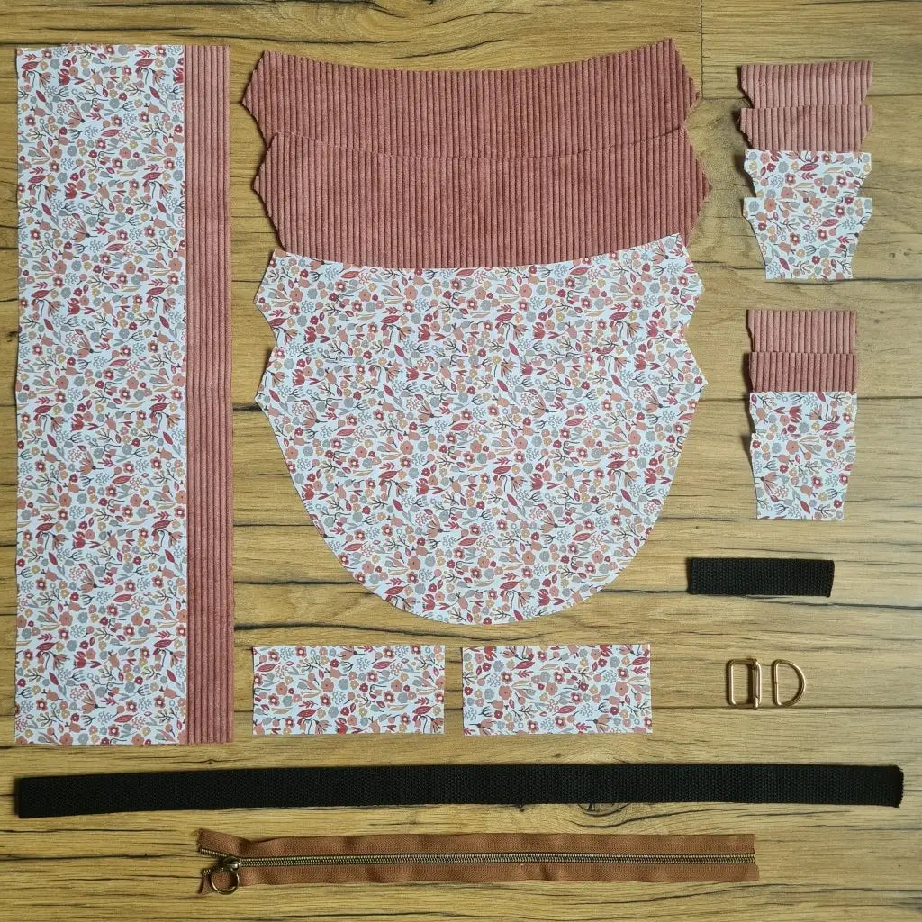 Kit "prêt à coudre" pour un sac demi-lune en velours côtelé au coloris rose avec une doublure en tissu à motif liberty. L'ensemble des pièces de tissu, déjà prédécoupées aux bonnes dimensions, sont disposées au sol et comprennent neuf pièces en tissu à motif liberty et sept pièces en velours côtelé de couleur rose ainsi qu'une fermeture éclair marron et une sangle noire.