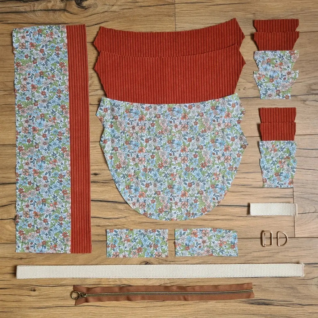 Kit "prêt à coudre" pour un sac demi-lune en velours côtelé au coloris terracotta avec une doublure en tissu à motif fleuri. L'ensemble des pièces de tissu, déjà prédécoupées aux bonnes dimensions, sont disposées au sol et comprennent neuf pièces en tissu à motif fleuri et sept pièces en velours côtelé de couleur terracotta ainsi qu'une fermeture éclair marron et une sangle beige.