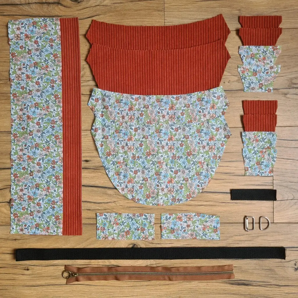 Kit "prêt à coudre" pour un sac demi-lune en velours côtelé au coloris terracotta avec une doublure en tissu à motif fleuri. L'ensemble des pièces de tissu, déjà prédécoupées aux bonnes dimensions, sont disposées au sol et comprennent neuf pièces en tissu à motif fleuri et sept pièces en velours côtelé de couleur terracotta ainsi qu'une fermeture éclair marron et une sangle noire.