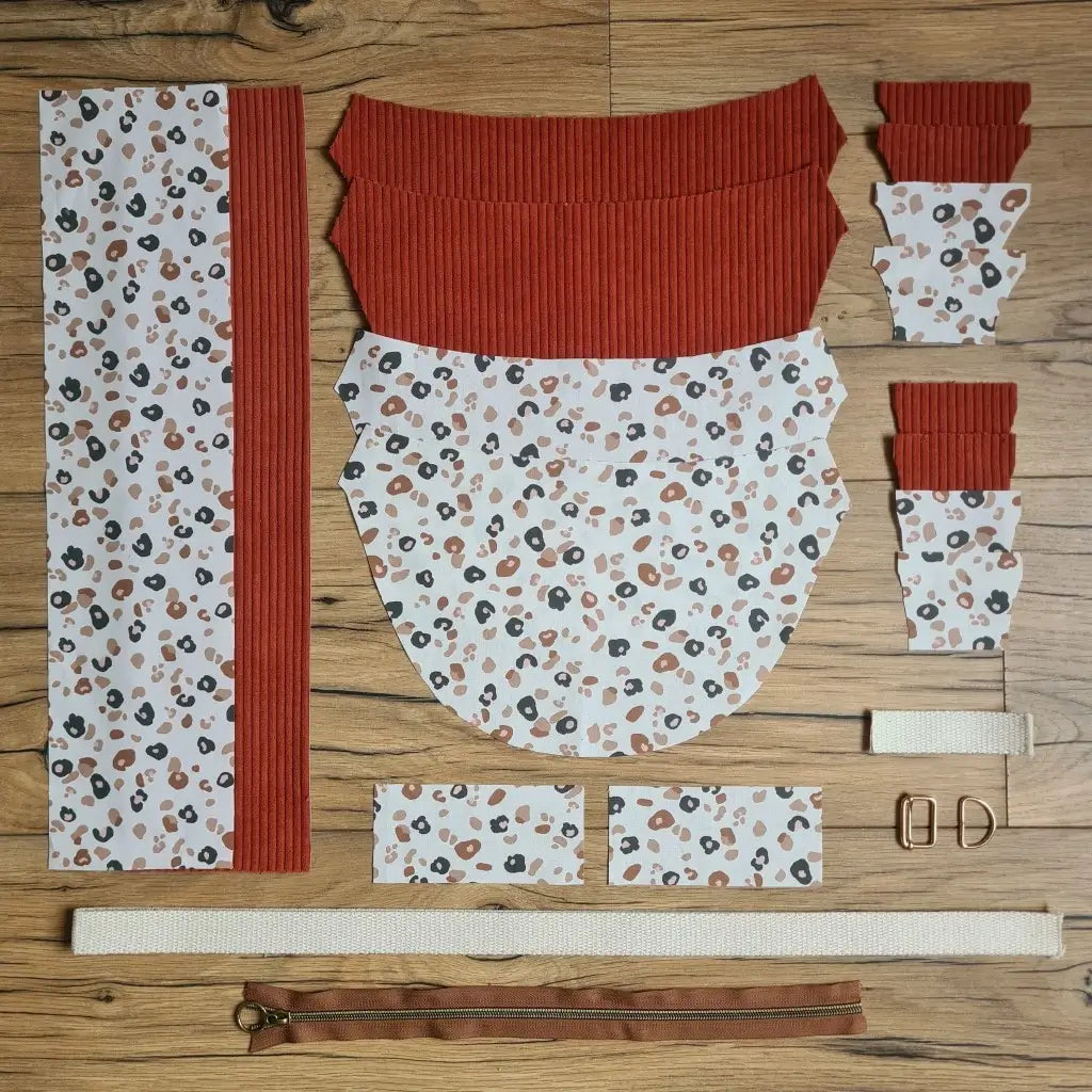 Kit "prêt à coudre" pour un sac demi-lune en velours côtelé au coloris terracotta avec une doublure en tissu à motif léopard. L'ensemble des pièces de tissu, déjà prédécoupées aux bonnes dimensions, sont disposées au sol et comprennent neuf pièces en tissu à motif léopard et sept pièces en velours côtelé de couleur terracotta ainsi qu'une fermeture éclair marron et une sangle beige.