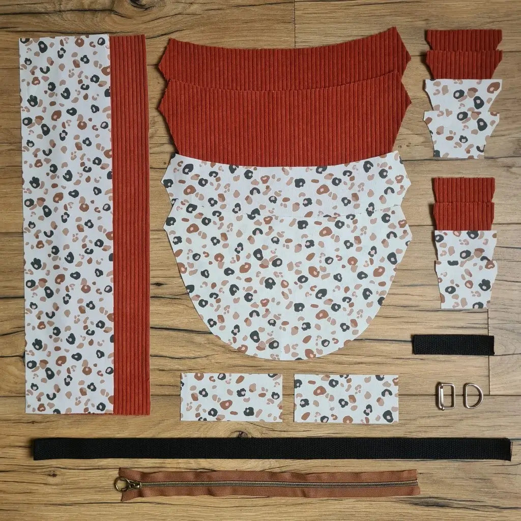 Kit "prêt à coudre" pour un sac demi-lune en velours côtelé au coloris terracotta avec une doublure en tissu à motif léopard. L'ensemble des pièces de tissu, déjà prédécoupées aux bonnes dimensions, sont disposées au sol et comprennent neuf pièces en tissu à motif léopard et sept pièces en velours côtelé de couleur terracotta ainsi qu'une fermeture éclair marron et une sangle noire.