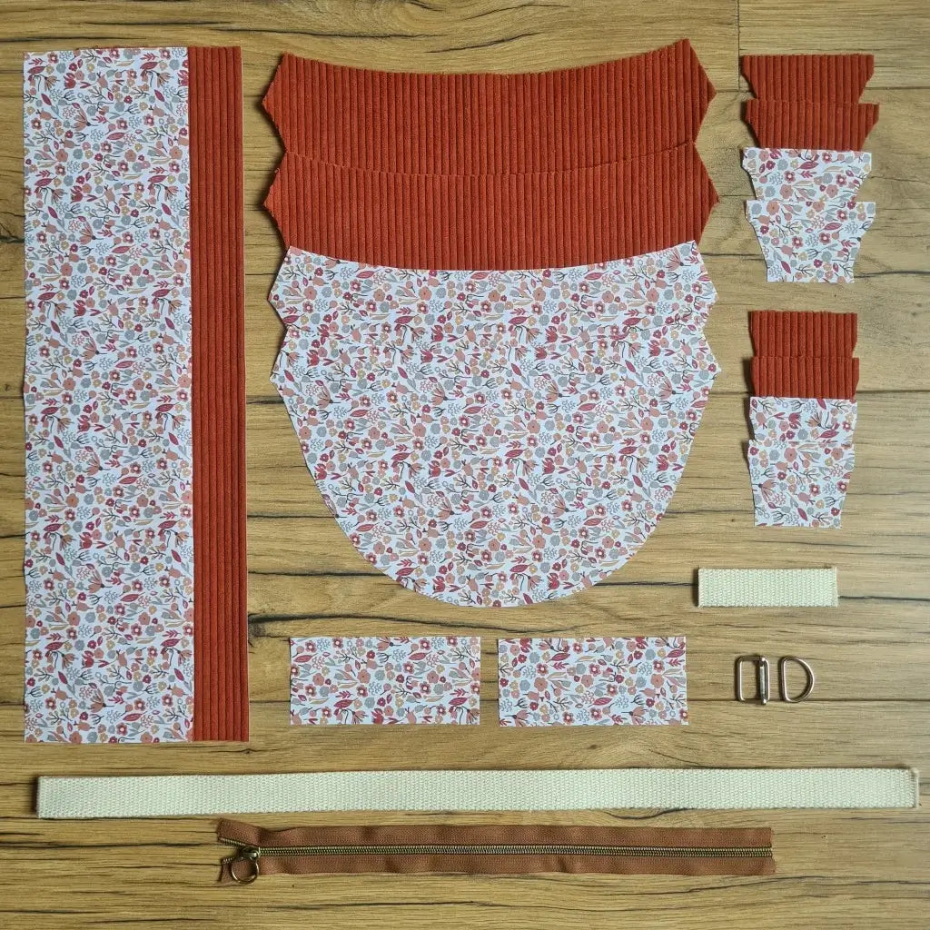 Kit "prêt à coudre" pour un sac demi-lune en velours côtelé au coloris terracotta avec une doublure en tissu à motif liberty. L'ensemble des pièces de tissu, déjà prédécoupées aux bonnes dimensions, sont disposées au sol et comprennent neuf pièces en tissu à motif liberty et sept pièces en velours côtelé de couleur terracotta ainsi qu'une fermeture éclair marron et une sangle beige.