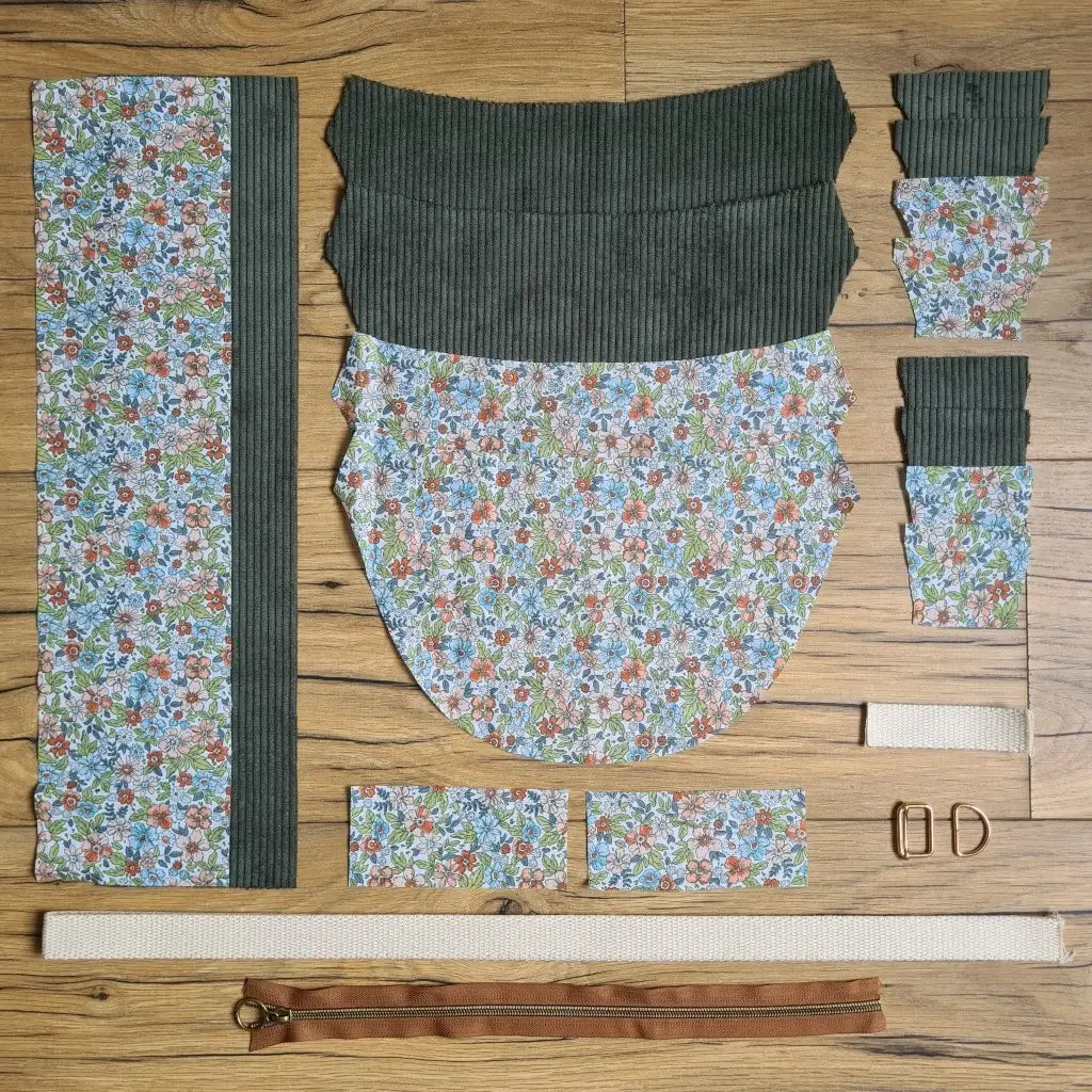 Kit "prêt à coudre" pour un sac demi-lune en velours côtelé au coloris vert avec une doublure en tissu à motif fleuri. L'ensemble des pièces de tissu, déjà prédécoupées aux bonnes dimensions, sont disposées au sol et comprennent neuf pièces en tissu à motif fleuri et sept pièces en velours côtelé de couleur verte ainsi qu'une fermeture éclair marron et une sangle beige.