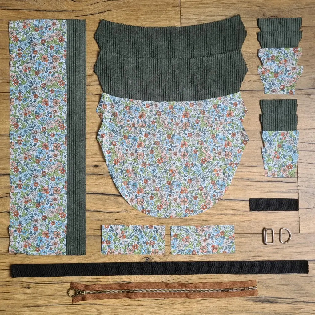 Kit "prêt à coudre" pour un sac demi-lune en velours côtelé au coloris vert avec une doublure en tissu à motif fleuri. L'ensemble des pièces de tissu, déjà prédécoupées aux bonnes dimensions, sont disposées au sol et comprennent neuf pièces en tissu à motif fleuri et sept pièces en velours côtelé de couleur verte ainsi qu'une fermeture éclair marron et une sangle noire.