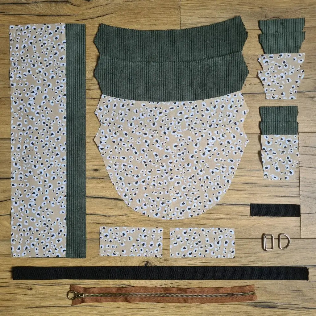 Kit "prêt à coudre" pour un sac demi-lune en velours côtelé au coloris vert avec une doublure en tissu à motif léopard. L'ensemble des pièces de tissu, déjà prédécoupées aux bonnes dimensions, sont disposées au sol et comprennent neuf pièces en tissu à motif léopard et sept pièces en velours côtelé de couleur verte ainsi qu'une fermeture éclair marron et une sangle noire.