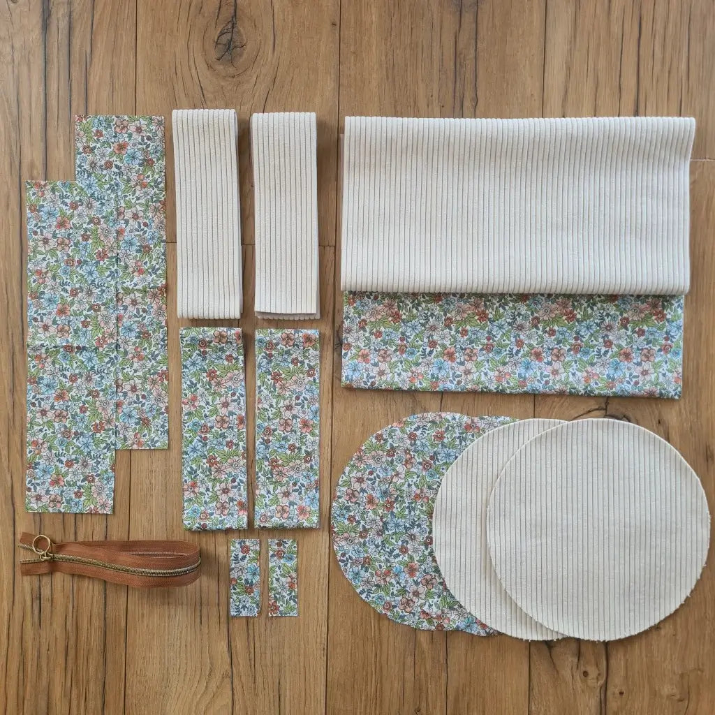 Kit "prêt à coudre" pour un sac polochon en velours côtelé au coloris beige avec une doublure en tissu à motif fleuri. L'ensemble des pièces de tissu, déjà prédécoupées aux bonnes dimensions, sont disposées au sol et comprennent neuf pièces en tissu à motif fleuri et cinq pièces en velours côtelé de couleur beige ainsi qu'une fermeture éclair marron.