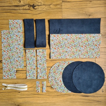 Kit "prêt à coudre" pour un sac polochon en velours côtelé au coloris bleu foncé avec une doublure en tissu à motif fleuri. L'ensemble des pièces de tissu, déjà prédécoupées aux bonnes dimensions, sont disposées au sol et comprennent neuf pièces en tissu à motif fleuri et cinq pièces en velours côtelé de couleur bleu foncé ainsi qu'une fermeture éclair blanche.