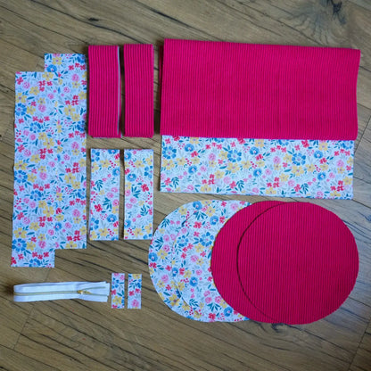 Kit prêt-à-coudre pour sac polochon composé de pièces de tissu prédécoupées : velours côtelé framboise et coton imprimé floral aux couleurs vives (jaune, bleu, rose, rouge). L’ensemble comprend également des parties rondes pour les extrémités, des bandes rectangulaires pour les anses et une fermeture éclair blanche avec curseur doré. Disposé à plat sur un sol en bois, ce kit complet permet de réaliser facilement un sac polochon artisanal pratique et coloré.