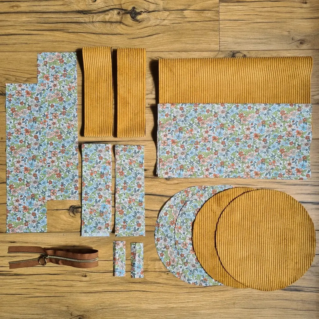 Kit "prêt à coudre" pour un sac polochon en velours côtelé au coloris jaune avec une doublure en tissu à motif fleuri. L'ensemble des pièces de tissu, déjà prédécoupées aux bonnes dimensions, sont disposées au sol et comprennent neuf pièces en tissu à motif fleuri et cinq pièces en velours côtelé de couleur jaune ainsi qu'une fermeture éclair marron.