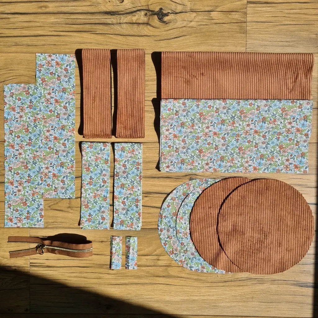 Kit "prêt à coudre" pour un sac polochon en velours côtelé au coloris marron avec une doublure en tissu à motif fleuri. L'ensemble des pièces de tissu, déjà prédécoupées aux bonnes dimensions, sont disposées au sol et comprennent neuf pièces en tissu à motif fleuri et cinq pièces en velours côtelé de couleur marron ainsi qu'une fermeture éclair marron.