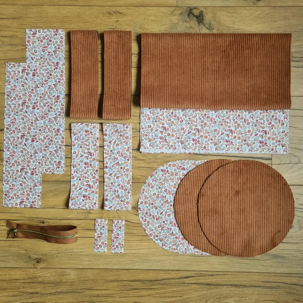 Kit "prêt à coudre" pour un sac polochon en velours côtelé au coloris marron avec une doublure en tissu à motif liberty. L'ensemble des pièces de tissu, déjà prédécoupées aux bonnes dimensions, sont disposées au sol et comprennent neuf pièces en tissu à motif liberty et cinq pièces en velours côtelé de couleur marron ainsi qu'une fermeture éclair marron.