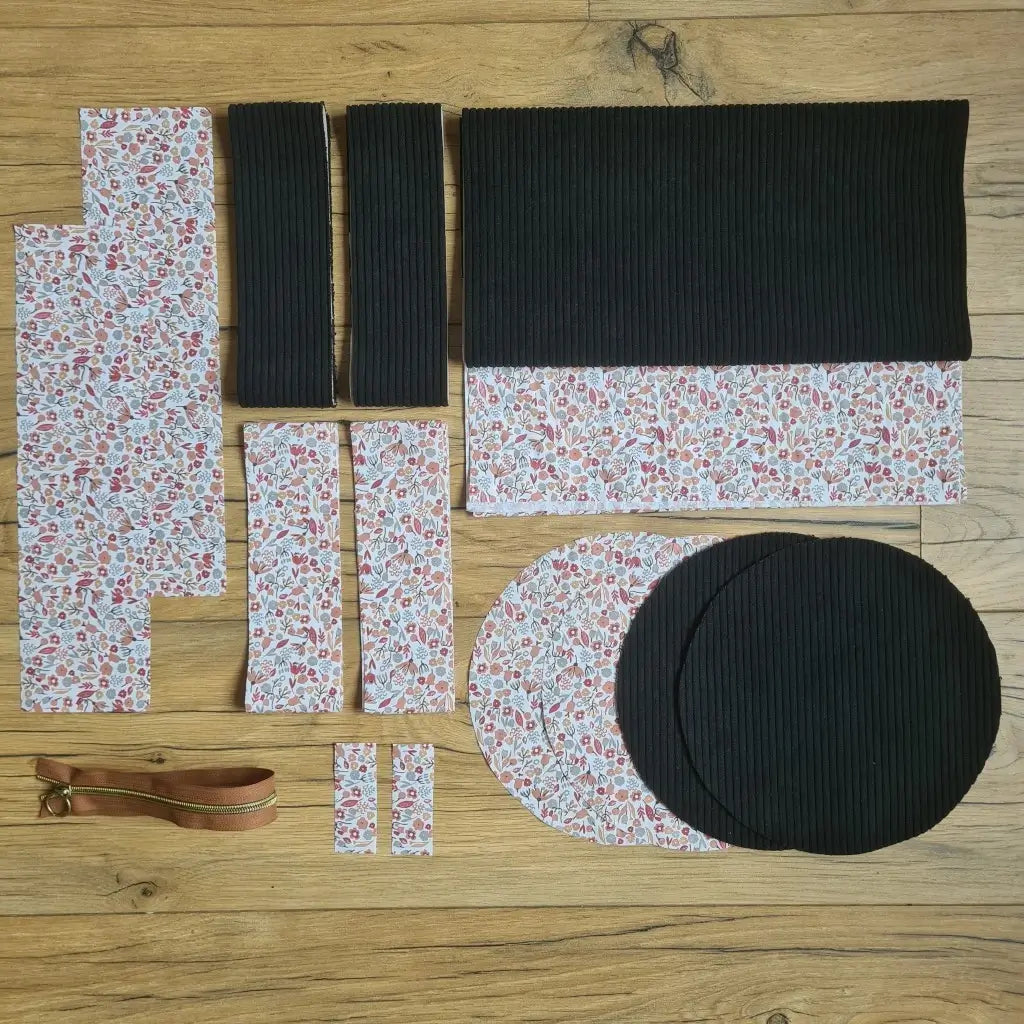 Kit "prêt à coudre" pour un sac polochon en velours côtelé au coloris noir avec une doublure en tissu à motif liberty. L'ensemble des pièces de tissu, déjà prédécoupées aux bonnes dimensions, sont disposées au sol et comprennent neuf pièces en tissu à motif liberty et cinq pièces en velours côtelé de couleur noir ainsi qu'une fermeture éclair marron.