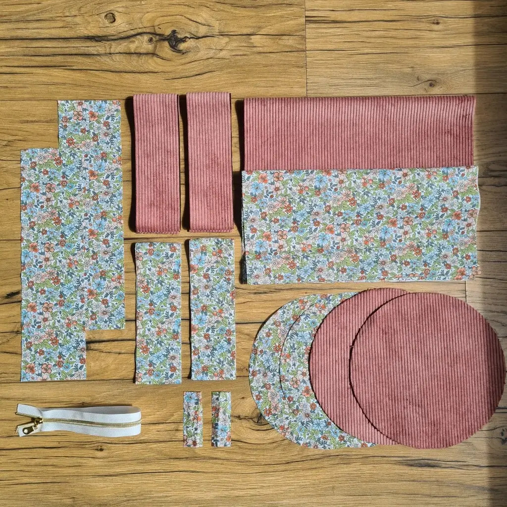 Kit "prêt à coudre" pour un sac polochon en velours côtelé au coloris rose avec une doublure en tissu à motif fleuri. L'ensemble des pièces de tissu, déjà prédécoupées aux bonnes dimensions, sont disposées au sol et comprennent neuf pièces en tissu à motif fleuri et cinq pièces en velours côtelé de couleur rose ainsi qu'une fermeture éclair blanche.