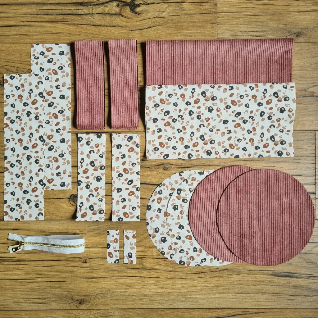 Kit "prêt à coudre" pour un sac polochon en velours côtelé au coloris rose avec une doublure en tissu à motif léopard. L'ensemble des pièces de tissu, déjà prédécoupées aux bonnes dimensions, sont disposées au sol et comprennent neuf pièces en tissu à motif léopard et cinq pièces en velours côtelé de couleur rose ainsi qu'une fermeture éclair blanche.