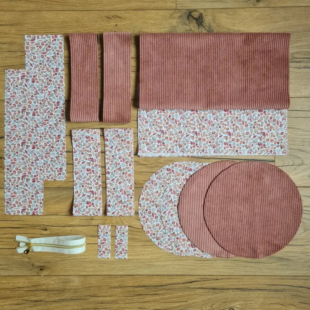 Kit "prêt à coudre" pour un sac polochon en velours côtelé au coloris rose avec une doublure en tissu à motif liberty. L'ensemble des pièces de tissu, déjà prédécoupées aux bonnes dimensions, sont disposées au sol et comprennent neuf pièces en tissu à motif liberty et cinq pièces en velours côtelé de couleur rose ainsi qu'une fermeture éclair blanche.