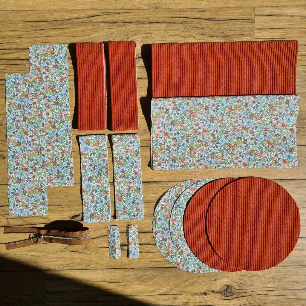 Kit "prêt à coudre" pour un sac polochon en velours côtelé au coloris terracotta avec une doublure en tissu à motif fleuri. L'ensemble des pièces de tissu, déjà prédécoupées aux bonnes dimensions, sont disposées au sol et comprennent neuf pièces en tissu à motif fleuri et cinq pièces en velours côtelé de couleur terracotta ainsi qu'une fermeture éclair marron.