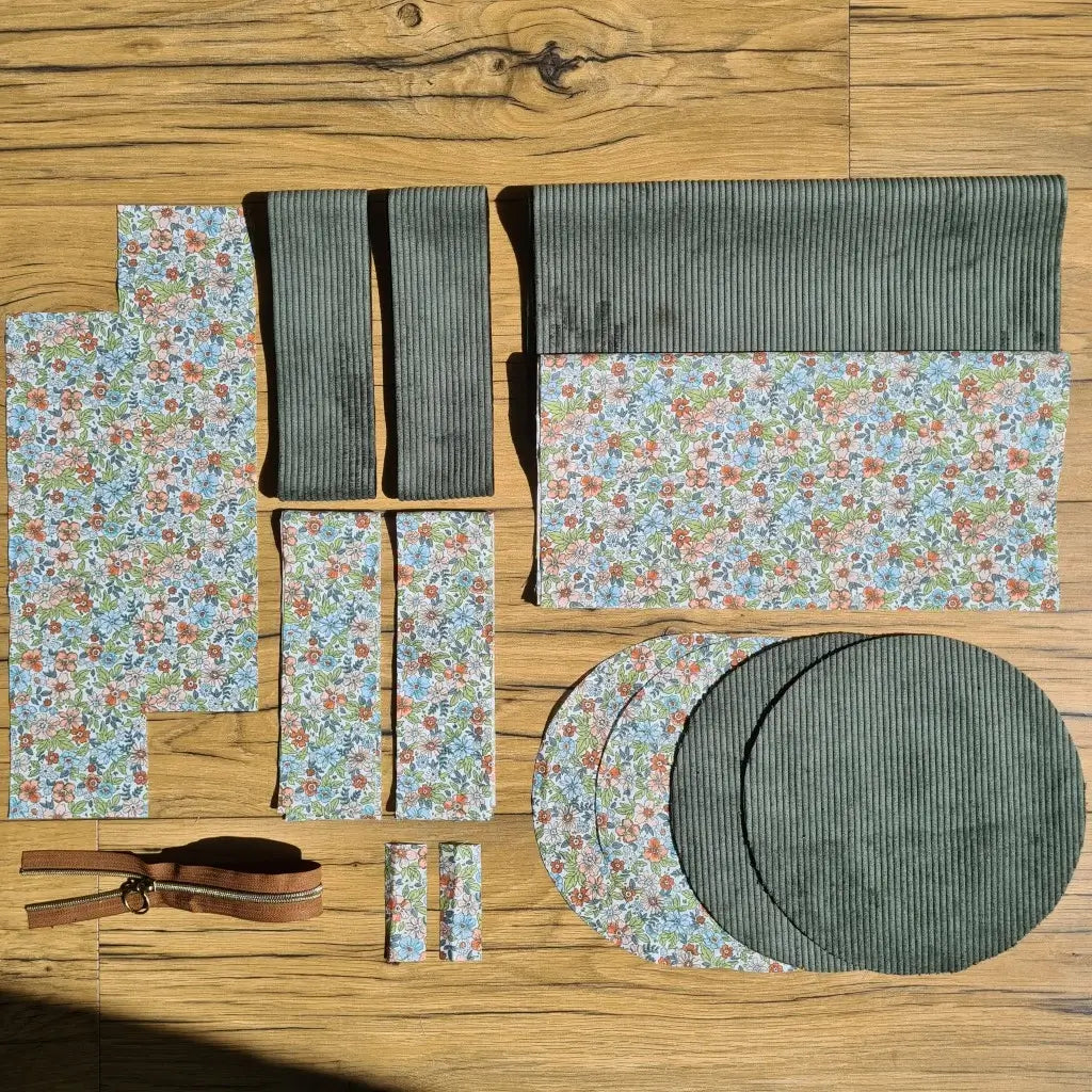 Kit "prêt à coudre" pour un sac polochon en velours côtelé au coloris vert avec une doublure en tissu à motif fleuri. L'ensemble des pièces de tissu, déjà prédécoupées aux bonnes dimensions, sont disposées au sol et comprennent neuf pièces en tissu à motif fleuri et cinq pièces en velours côtelé de couleur verte ainsi qu'une fermeture éclair marron.