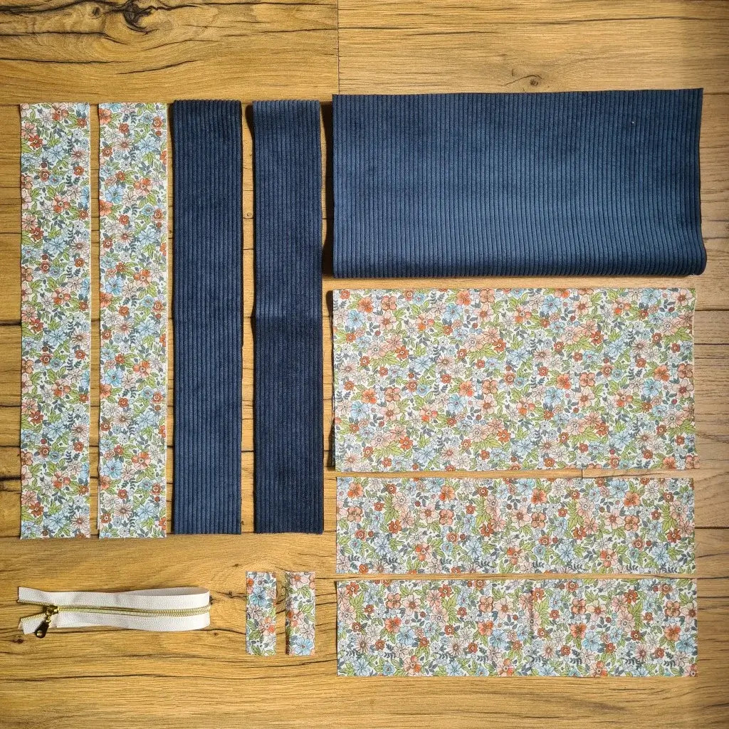 Kit "prêt à coudre" pour un tote-bag en velours côtelé au coloris bleu foncé avec une doublure en tissu à motif fleuri. L'ensemble des pièces de tissu, déjà prédécoupées aux bonnes dimensions, sont disposées au sol et comprennent sept pièces en tissu à motif fleuri et trois pièces en velours côtelé de couleur bleu foncé ainsi qu'une fermeture éclair blanche.