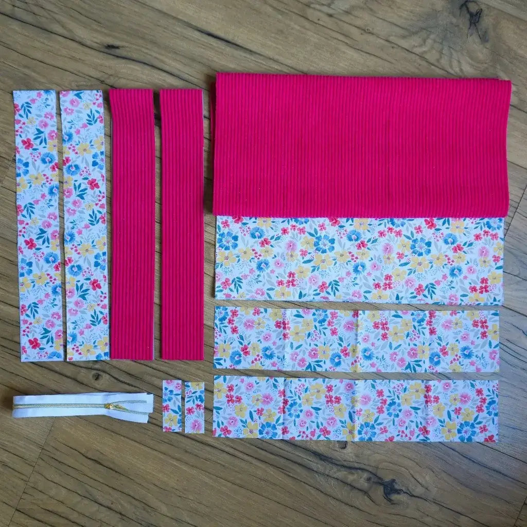 Kit prêt-à-coudre pour tote bag, comprenant des pièces de tissu prédécoupées en velours côtelé framboise et en coton imprimé floral multicolore (jaune, bleu, rouge, rose). Le lot inclut également des bandes pour les anses, un morceau pour la base et une fermeture éclair blanche à curseur doré. Présenté à plat sur un sol en bois, ce kit complet permet de réaliser un sac cabas artisanal pratique et coloré, idéal pour une création couture personnalisée.
