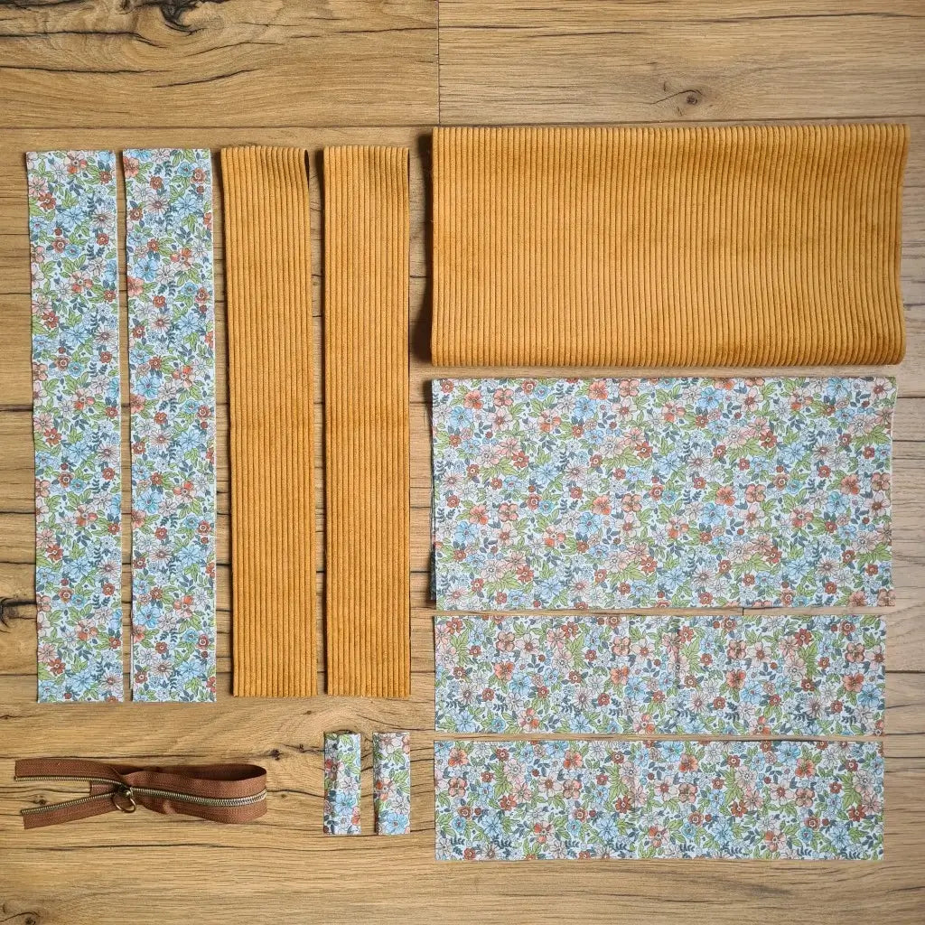 Kit "prêt à coudre" pour un tote-bag en velours côtelé au coloris jaune avec une doublure en tissu à motif fleuri. L'ensemble des pièces de tissu, déjà prédécoupées aux bonnes dimensions, sont disposées au sol et comprennent sept pièces en tissu à motif fleuri et trois pièces en velours côtelé de couleur jaune ainsi qu'une fermeture éclair marron.