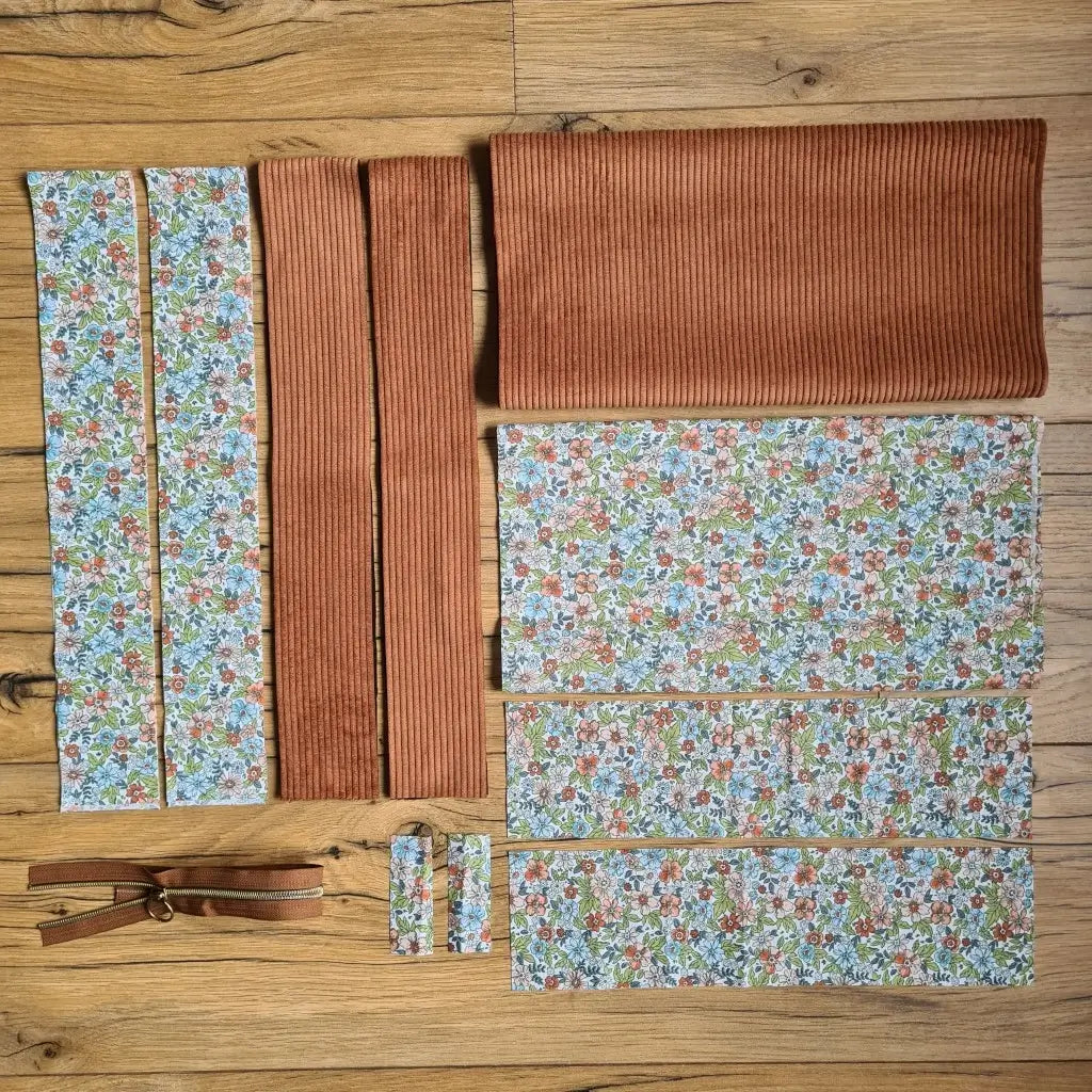 Kit "prêt à coudre" pour un tote-bag en velours côtelé au coloris marron avec une doublure en tissu à motif fleuri. L'ensemble des pièces de tissu, déjà prédécoupées aux bonnes dimensions, sont disposées au sol et comprennent sept pièces en tissu à motif fleuri et trois pièces en velours côtelé de couleur marron ainsi qu'une fermeture éclair marron.