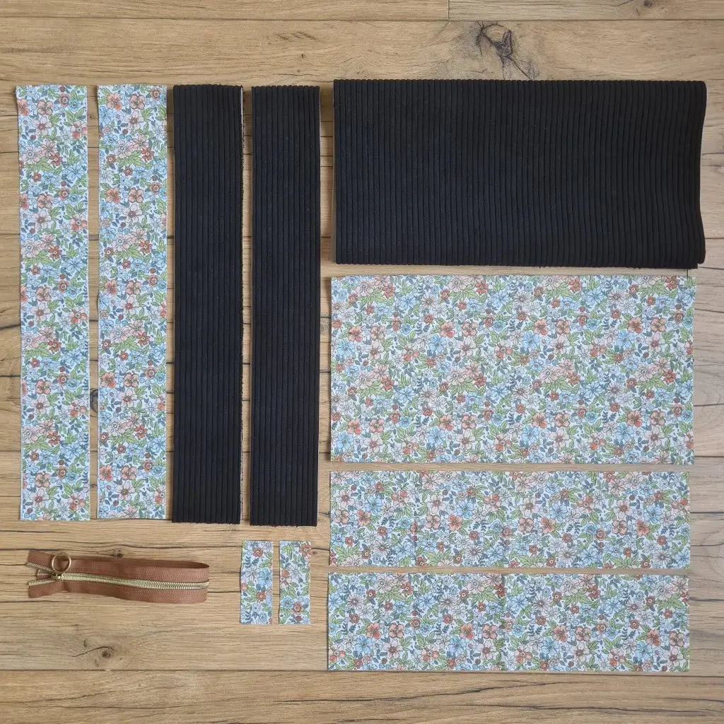 Kit "prêt à coudre" pour un tote-bag en velours côtelé au coloris noir avec une doublure en tissu à motif fleuri. L'ensemble des pièces de tissu, déjà prédécoupées aux bonnes dimensions, sont disposées au sol et comprennent sept pièces en tissu à motif fleuri et trois pièces en velours côtelé de couleur noir ainsi qu'une fermeture éclair marron.