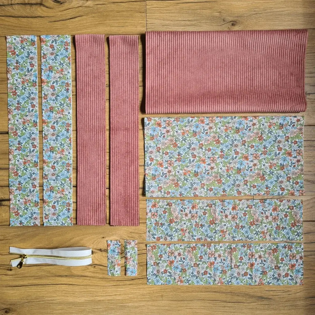 Kit "prêt à coudre" pour un tote-bag en velours côtelé au coloris rose avec une doublure en tissu à motif fleuri. L'ensemble des pièces de tissu, déjà prédécoupées aux bonnes dimensions, sont disposées au sol et comprennent sept pièces en tissu à motif fleuri et trois pièces en velours côtelé de couleur rose ainsi qu'une fermeture éclair blanche.