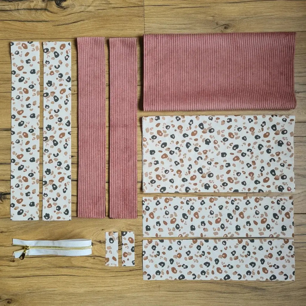 Kit "prêt à coudre" pour un tote-bag en velours côtelé au coloris rose avec une doublure en tissu à motif léopard. L'ensemble des pièces de tissu, déjà prédécoupées aux bonnes dimensions, sont disposées au sol et comprennent sept pièces en tissu à motif léopard et trois pièces en velours côtelé de couleur rose ainsi qu'une fermeture éclair blanche.