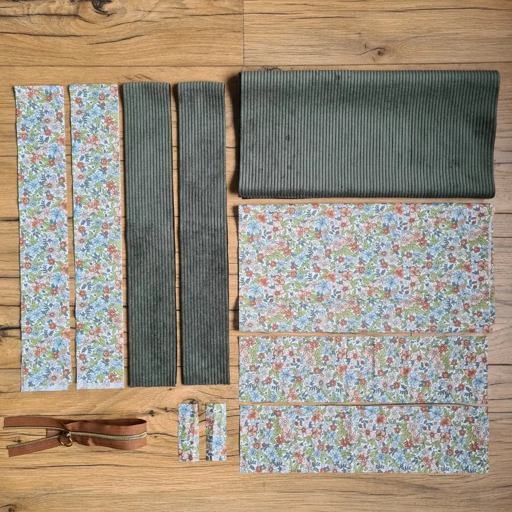 Kit "prêt à coudre" pour un tote-bag en velours côtelé au coloris vert avec une doublure en tissu à motif fleuri. L'ensemble des pièces de tissu, déjà prédécoupées aux bonnes dimensions, sont disposées au sol et comprennent sept pièces en tissu à motif fleuri et trois pièces en velours côtelé de couleur verte ainsi qu'une fermeture éclair marron.